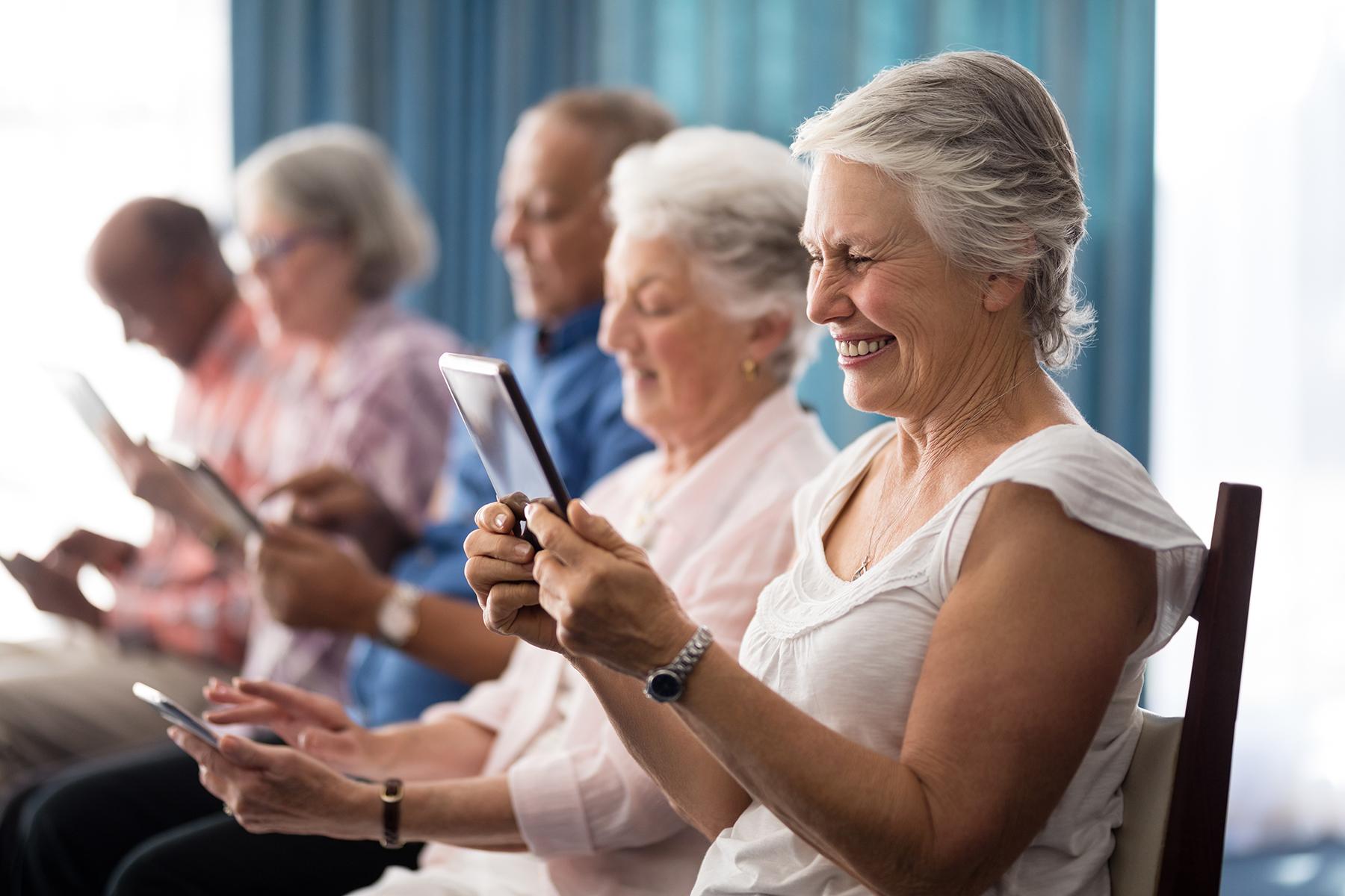 Row of smiling senior people sitting on chairs using digital tablets at retirement home